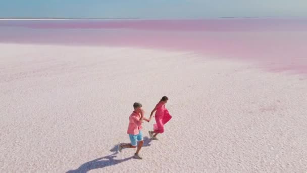 Imágenes aéreas de drones de feliz pareja joven en ropa rosa que se divierten y corren alegremente en la costa blanca del colorido lago mineral rosa brillante — Vídeo de stock