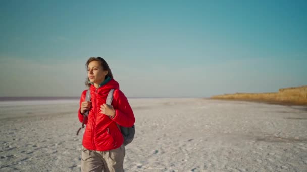 Slow motion portret van mooie zelfverzekerde en rustige jonge vrouw wandelaar in rode jas wandelen op zoute meerkust — Stockvideo