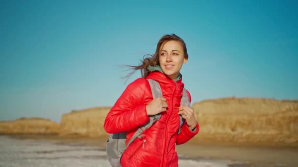 Zeitlupenporträt einer fröhlich lächelnden und fröhlichen jungen Wanderin in roter Jacke, die am salzigen Seeufer spaziert — Stockvideo