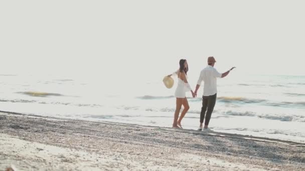 Романтическая пара в шляпах, идущая вдоль песчаного пляжа, держась за руки. — стоковое видео