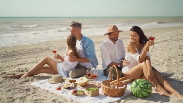 Cuatro amigos milenarios tienen picnic en la playa de arena en el día soleado siguiente mar ondulante — Vídeo de stock