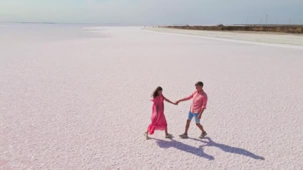 Luchtfoto drone beelden van gelukkig jong paar in roze slijtage hebben plezier en vrolijk lopen op witte kust van heldere kleurrijke roze mineraal meer — Stockvideo