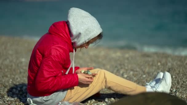 Young woman in red jacket and hoodie sitting on pebble sea beach at windy sunny day and picking up pebble stones — Stock Video