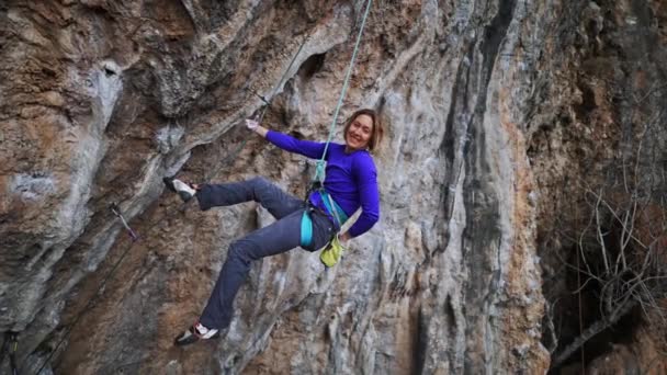 Cámara lenta sonriendo mujer cansada escalador de roca colgando de la cuerda en la ruta de desafío duro en el acantilado colgante, descansando y tiza manos — Vídeo de stock