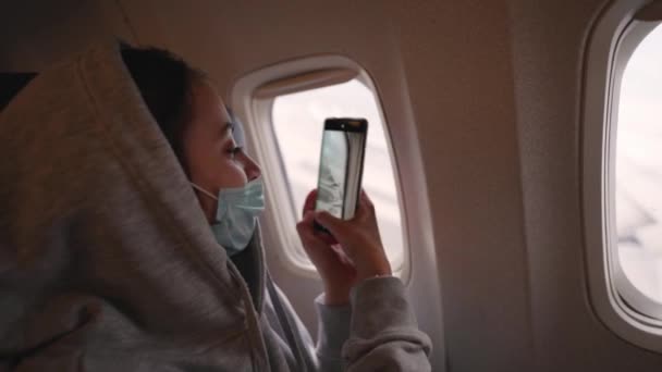 Boldog női turista orvosi maszk nézi repülőgép ablak repülőgép kabin és forgatás mobiltelefonnal. — Stock videók