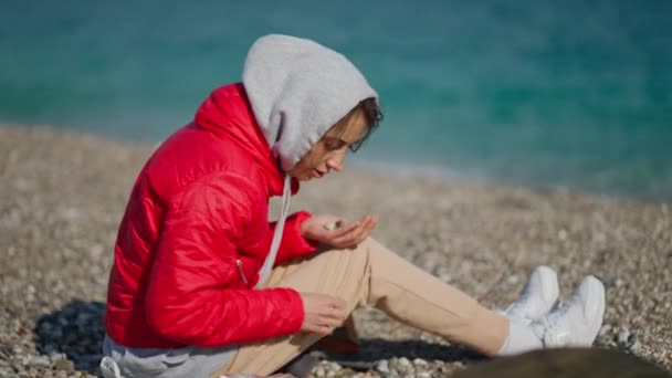 Jovem mulher em casaco vermelho e capuz sentado na praia do mar de seixos no dia ensolarado ventoso e pegando pedras de seixos — Vídeo de Stock