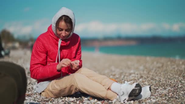 Jonge vrouw in rode jas en hoodie zitten op kiezelstrand op winderige zonnige dag en het oppakken van kiezelstenen — Stockvideo
