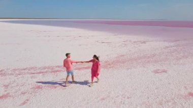 Pembe elbiseli mutlu çiftin parlak pembe mineral gölünün beyaz kıyısında eğlenirken ve neşe içinde koşarken çekilmiş hava aracı görüntüleri.