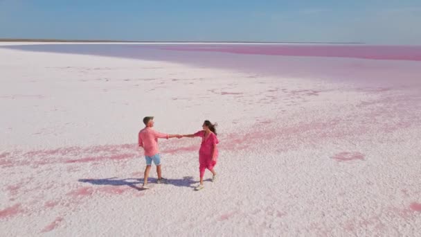 Imagens aéreas drone de casal feliz jovem em desgaste rosa se divertindo e alegremente correndo na costa branca do lago mineral rosa colorido brilhante — Vídeo de Stock