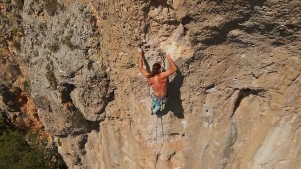 Aerial slow motion filming from drone strong muscular man rock climber climbs on vertical cliff and makes hard efforts, gripping holds. — стоковое видео