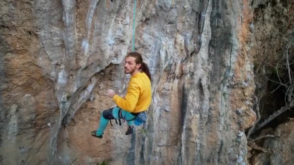 Zeitlupe eines fröhlichen, gut aussehenden Bergsteigers in gelbem Sweatshirt mit langen Haaren, die am Seil hängen und nach erfolgreicher Besteigung der Route auf einer Klippe hoch fünf geben. — Stockvideo