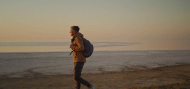 Sırt çantalı bir turist kıyı şeridinde yürüyor, altın gündoğumuyla sabah buluşuyor.