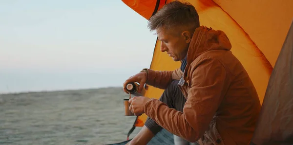Mies matkustava istuu leiriteltassa ja kaatamalla lämmintä kahvia termospullo muki — kuvapankkivalokuva