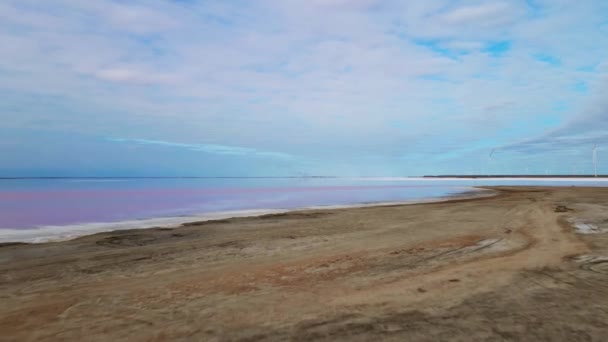 Vol de drone sur la surface lisse du lac rose coloré au lever du soleil nuageux avec parc éolien sur fond. — Video