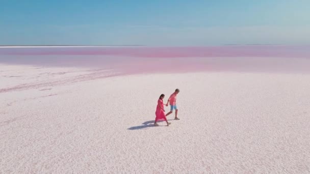 Imágenes aéreas de drones de feliz pareja joven en ropa rosa que se divierten y caminan alegremente en la costa blanca del colorido lago mineral rosa brillante — Vídeos de Stock