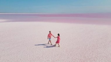 Pembe elbiseli mutlu çiftin hava aracı görüntüleri. Parlak pembe mineral gölünün beyaz kıyısında neşeyle yürüyor ve eğleniyorlar.