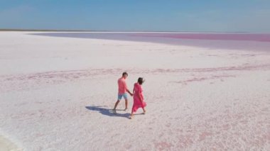 Pembe elbiseli mutlu çiftin parlak pembe mineral gölünün beyaz kıyısında eğlenirken ve neşe içinde koşarken çekilmiş hava aracı görüntüleri.