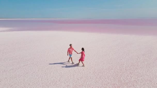 Luchtfoto drone beelden van gelukkig jong paar in roze dragen plezier hebben en vrolijk wandelen op witte kust van heldere kleurrijke roze mineraal meer — Stockvideo