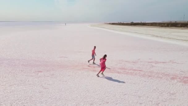 Съемка с беспилотника счастливой молодой пары в розовой одежде, веселящейся и радостно бегущей по белому побережью ярко-красочного розового минерального озера — стоковое видео