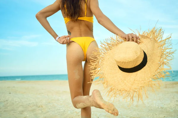 Bronzeada mulher de corpo magro em biquíni sexy andando na praia de areia, segurando chapéu de palha. — Fotografia de Stock