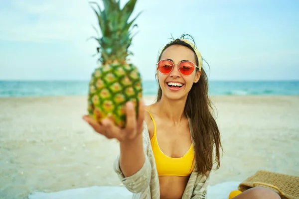 Smilende jente i røde solbriller som ser på kamera og holder ananas på stranden – stockfoto