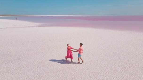 Luchtfoto van een gelukkig romantisch paar dat handen in handen loopt en kust. man en vrouw in roze dragen wandelingen op witte kust van heldere kleurrijke meer met roze mineraalwater — Stockvideo