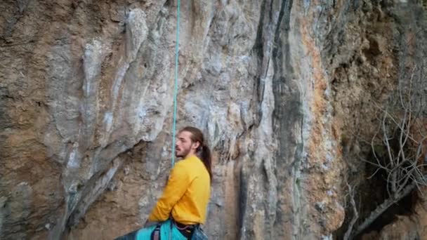 Cámara lenta de alegre hombre guapo escalador de roca en sudadera amarilla con el pelo largo colgando de la cuerda y dar cinco altos después de la ascensión exitosa de la ruta en el acantilado. — Vídeo de stock