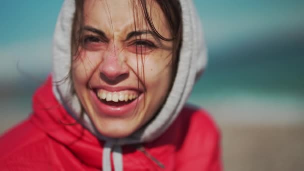 Primer plano retrato de joven feliz sonriente mujer en chaqueta roja y sudadera con capucha sentado en la playa de guijarros del mar en el día soleado ventoso. — Vídeos de Stock