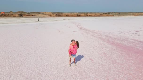 Cirkel drone vlucht en luchtfoto van gelukkig romantisch mooi paar in roze slijtage staande op witte zoute kust van minerale meer. man houdt vrouw op handen en kussen. — Stockvideo