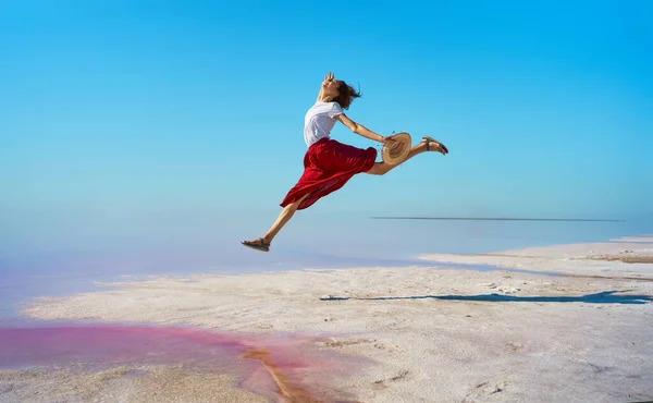 Lykkelig kvinne i rødt skjørt hopper, nyter rosa saltsjø med blå himmel – stockfoto