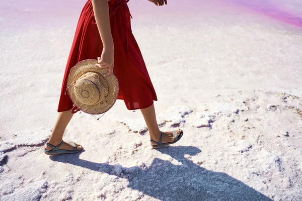 Naisen jalat punaisessa hameessa kävely suolalla valkoisella rannalla, pitäen olkihattua — kuvapankkivalokuva
