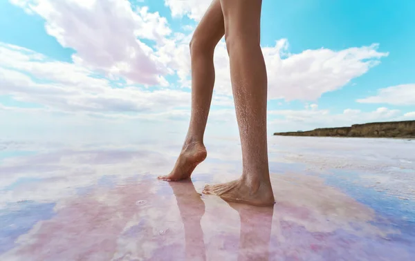 Piernas largas femeninas con sal en la piel, de pie en el lago de color rosa sal, reflejo en el agua del cielo nublado — Foto de Stock