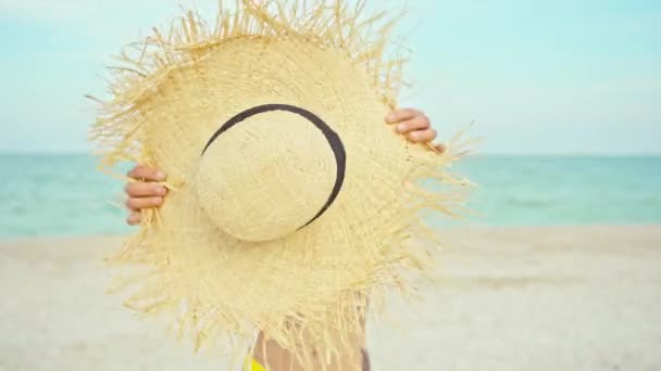 Mujer sosteniendo sombrero de paja de verano delante de la cámara en la playa del océano ventoso — Vídeos de Stock