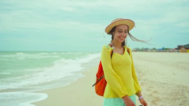 Radosna kobieta w kapeluszu w żółtej koszuli bawiąca się i czująca szczęście na plaży z falami — Wideo stockowe