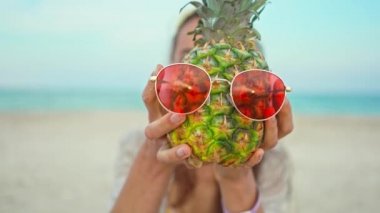 Sahilde elinde kırmızı güneş gözlüklü meyve ananasıyla gülen bir kadın.