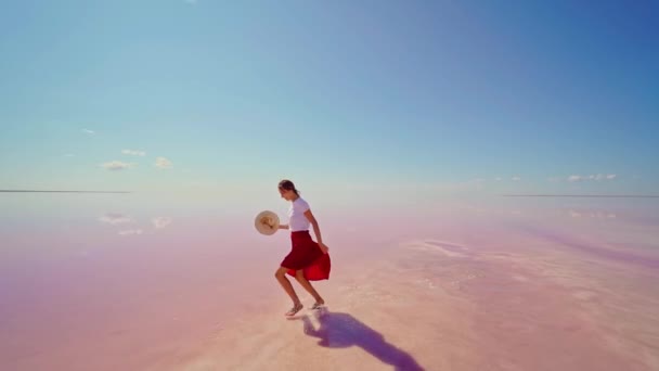 Ευτυχισμένη γυναίκα με κόκκινη φούστα τρέχει, απολαμβάνοντας φωτεινό ροζ αλάτι λίμνη με μπλε ουρανό — Αρχείο Βίντεο