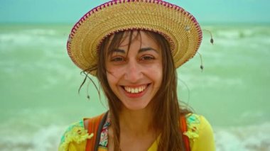 Kamera yakın plan portresinden fırlıyor. Şapkalı mutlu genç kadın deniz sahilinde tatil yapıyor. Rüzgarlı bir günde dalgalarla kendini iyi hissediyor.