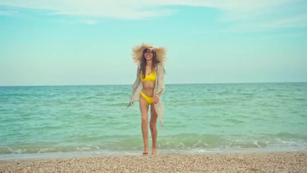 Radosny szczęśliwy podróżnik kobieta w modne stroje kąpielowe i słomkowy kapelusz idzie na kamery wiht widok na morze na tle. — Wideo stockowe