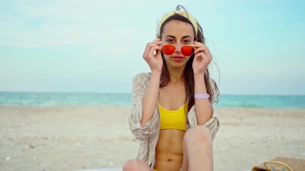 Primer plano retrato de moda al aire libre mujer joven con bikini amarillo y gafas de sol de color rosa por mar en la playa tropical — Vídeos de Stock