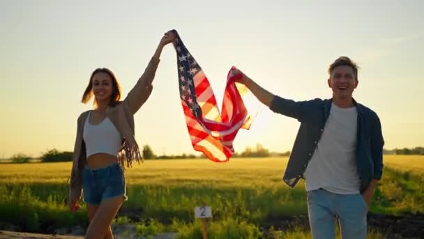 Gelukkig glimlachend man en vrouw, amerikaanse familie, wandelen in het veld bij zonsondergang met vlag van de VS met trots, viering 4 juli — Stockvideo