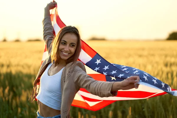 Jovem mulher feliz acenando EUA estrelas e listras bandeira no pôr do sol dourado campo de sol — Fotografia de Stock