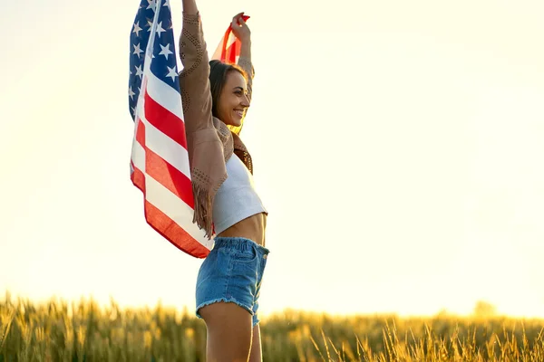 Donna deliziata in piedi al campo al tramonto con le mani alzate, tenendo bandiera nazionale americana — Foto Stock