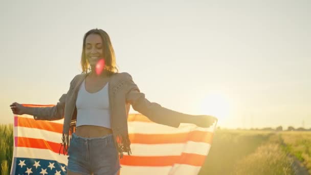 Självsäker kvinna viftar med stjärnor och ränder USA flagga på fältet vid solnedgången. Självständighetsdagen — Stockvideo