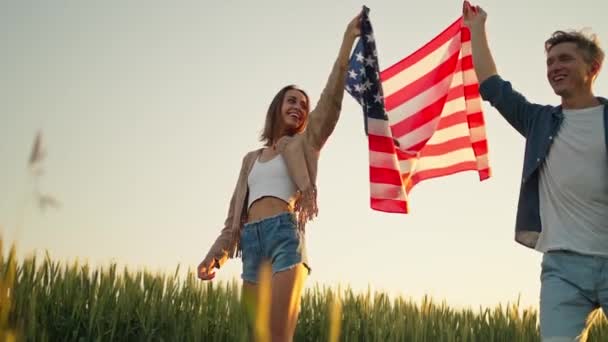 4k câmera lenta de feliz casal patriótico celebrando o quarto de julho com bandeira nacional em lardmark rural ao pôr do sol — Vídeo de Stock