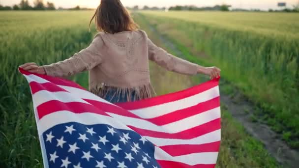 Mladá šťastná usměvavá dívka běží s vlajkou USA nad pšeničným polem a otočí se — Stock video