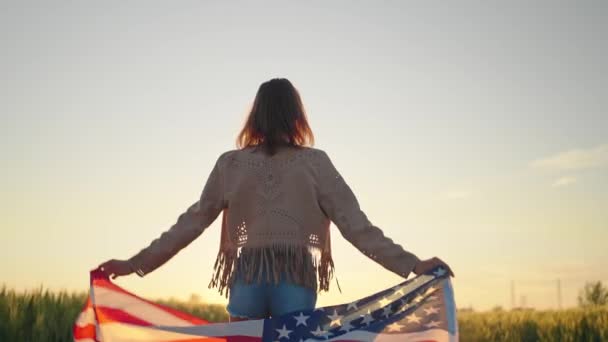 Câmera lenta visão traseira da jovem bela mulher americana sexy segurando uma bandeira americana caminhando campo de trigo ao pôr do sol — Vídeo de Stock