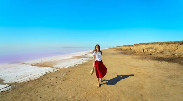 Kvinne med rødt skjørt som går på sandstrand med blå himmel nær salt rosa innsjø – stockfoto