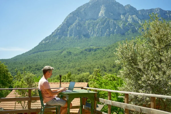 Mies freelancer työskentelee kannettavan tietokoneen terassilla vuoristomaisemilla. Teknologia ja matkustaminen — kuvapankkivalokuva