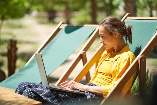 Mujer freelance calificada usando computadora portátil mientras está sentado en la tumbona al aire libre, digital de trabajo en línea —  Fotos de Stock