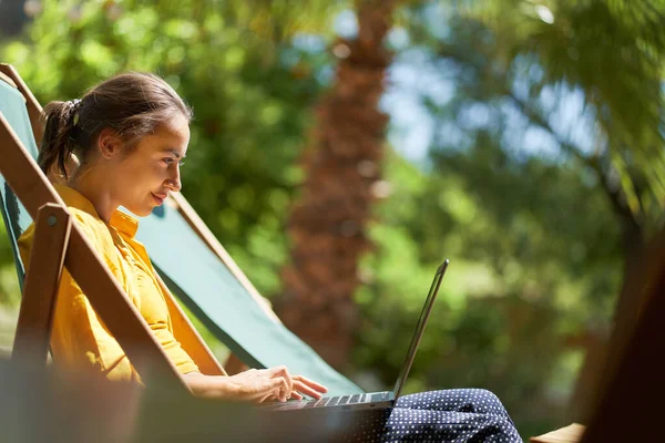 Side view kauko verkossa työskentelevä nainen istuu aurinkotuolissa, kannettavan tietokoneen avulla — kuvapankkivalokuva